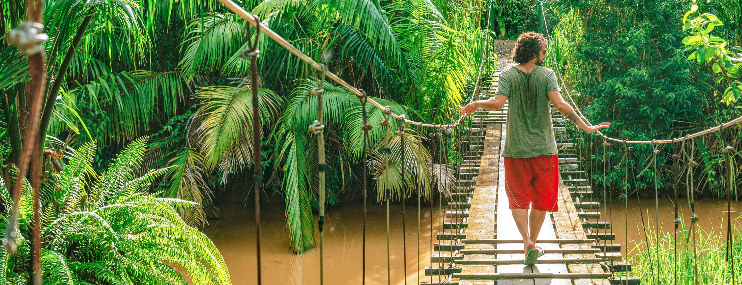 Homme marchant sur un pont en bois au-dessus d'une rivière, entouré de végétation luxuriante.