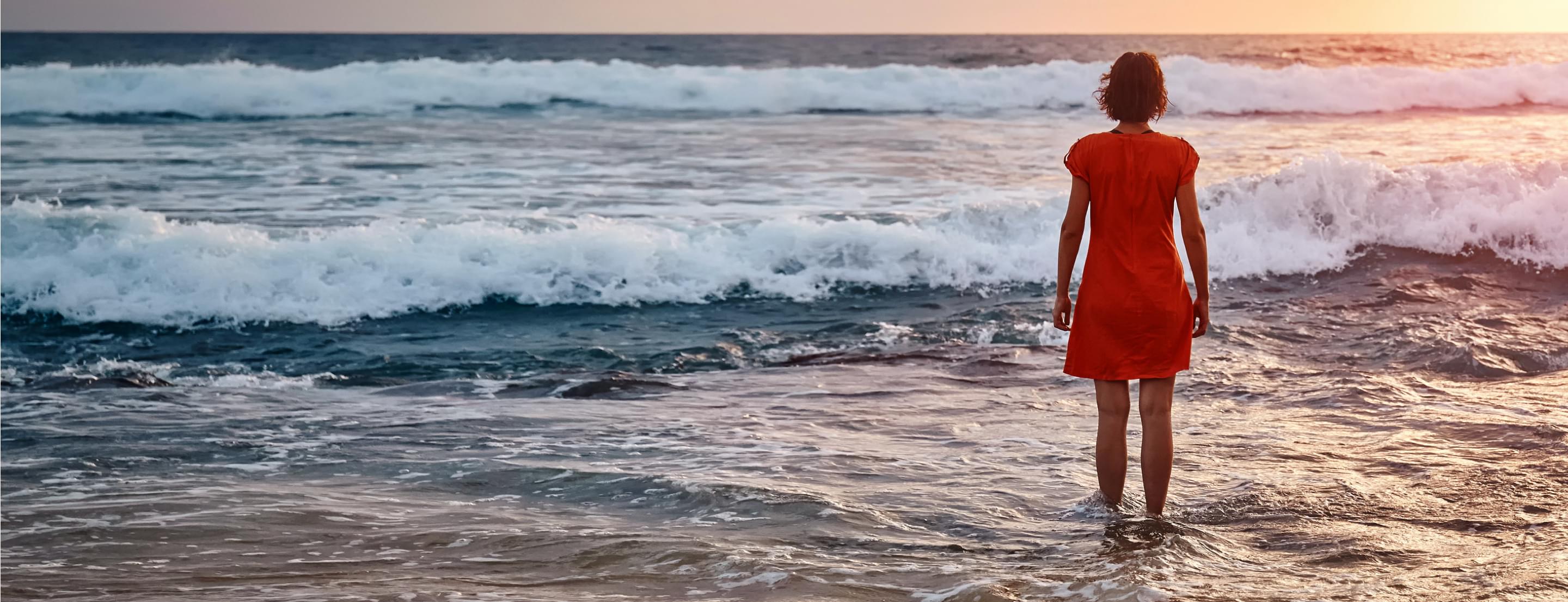 Femme en robe rouge se tenant dans l'eau, regardant les vagues sous un ciel au coucher de soleil.