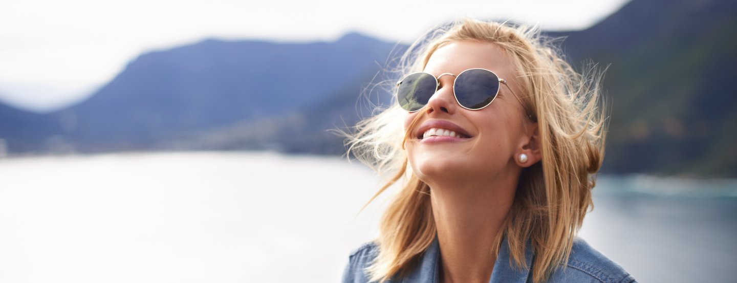 Visage d'une femme aux cheveux blonds, portant des lunettes de soleil, devant un lac de montagne