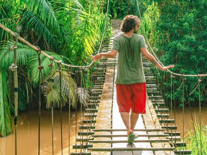Homme marchant sur un pont en bois au-dessus d'une rivière, entouré de végétation luxuriante.