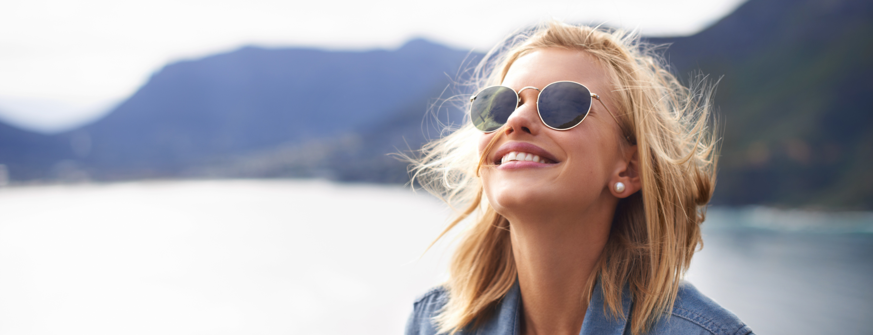 Visage d'une femme aux cheveux blonds, portant des lunettes de soleil, devant un lac de montagne