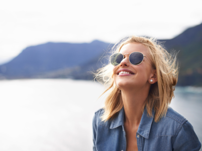 Une femme aux cheveux blonds, portant des lunettes profite du soleil, devant un lac de montagne